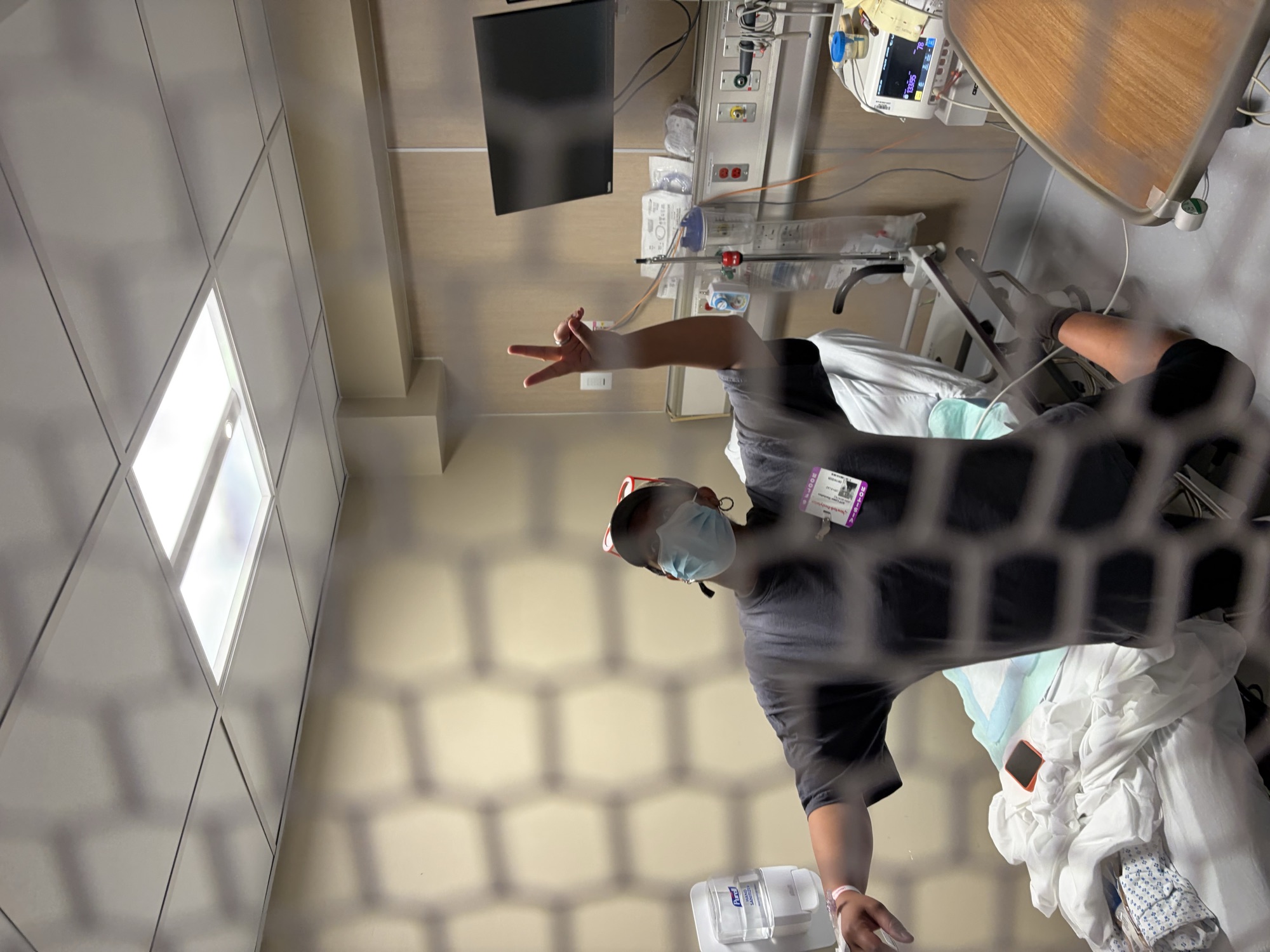 Jara giving a peace sign in her hospital room, hospital bed and monitoring equipment visible behind her