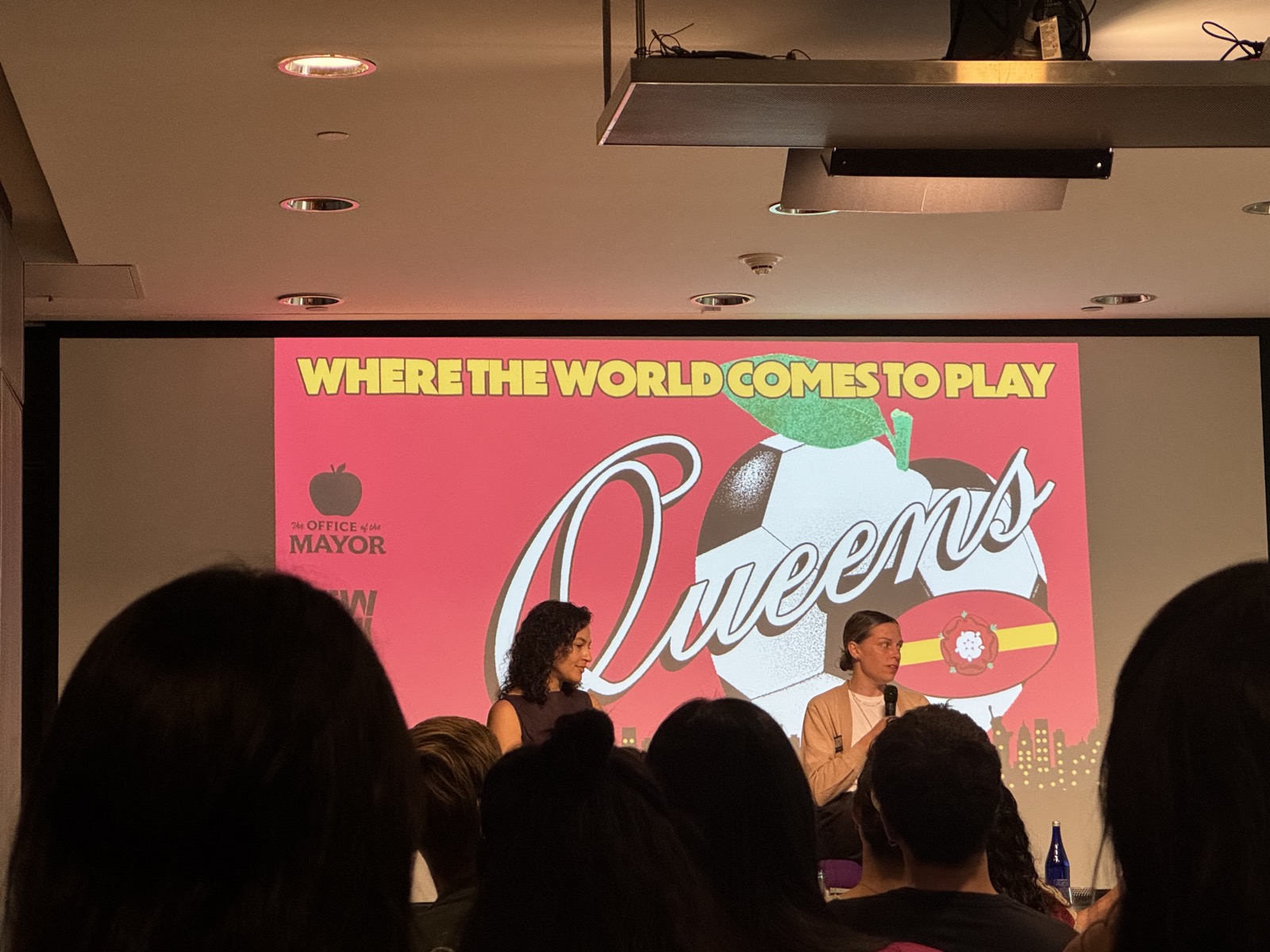 Screen reading Where the World Comes to Play Queens, with two panelists seated on stage
